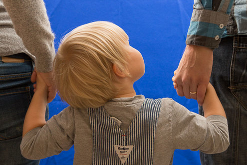 Lapsi seisoo kahden aikuisen välissä ja pitää heitä käsistä kiinni.
