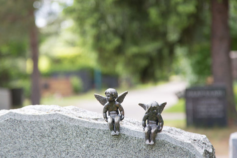 Kaksi raudasta valettua enkelilasta istuvat graniittisella hautakivellä katselleen ympärilleen. Taustalla näkyy hautausmaan vehreys.