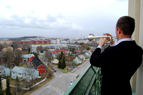 Mies seisoo selin ja soittaa trumpettia. Taustalla on kaupunkimaisema, jossa on lähinnä rintamamiestaloja.