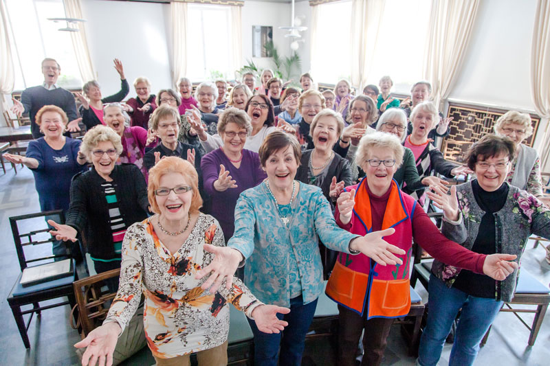 Värikäisiin vaatteisiin pukeutunut naisjoukko seisoo ryhmässä kädet avattuena eteen alaviistoon.