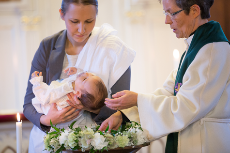 Nainen pitäää vauvaa sylissään ja heidän vasemmalla puolella oleva pappi valuttaa vettä vauvan päähän.