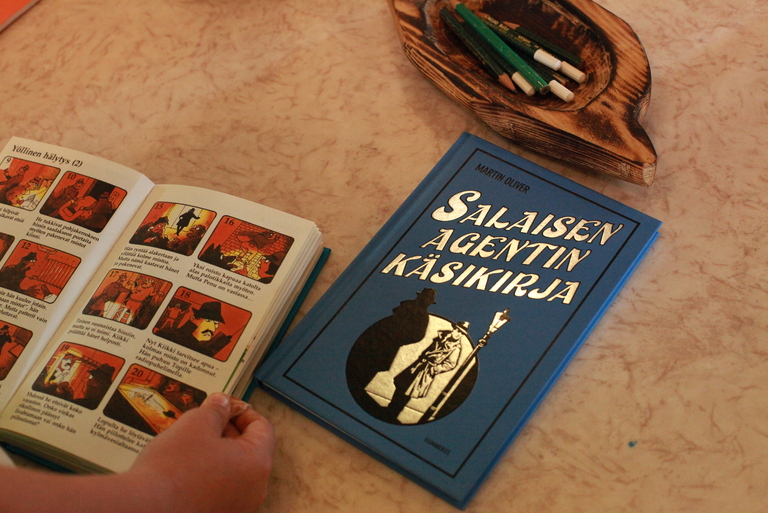 Pöydällä on Salaisen Agentikäsikirjan kansija avonainen salapoliisisarjakuva.