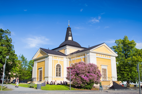 Tampereen keskustorin Vanha kirkko pilvettömänä kesäpäivänä. Ihmisiä istuskelee kirkon edustalla nurmella kiireettömästi.