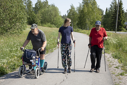 Kaksi miestä ja keskellä nainen kulkevat kävelytietä. Nainen ja toinen mies sauvakävelevät ja toinen mies potkii potku-rollaattorilla.