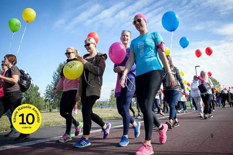 Etualalla neljä naista kävelemässä värikkäissä urheiluvaatteissa ja ilmapallot käsissä. Taustalla tulee iso kävelijäjoukko.