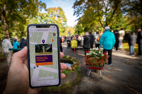 Puhelimen näytöllö Kalevankangas.fi-sivusto. Taustalla ihmisiä kulkemassa Kalevankaan hautausmaalla.