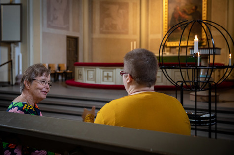 Päivystäjä keskustelee seurakuntalaisen kanssa kirkonpenkissä.