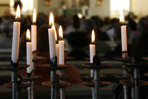 Kynttelikön, jossa on palavat kynttilän, takana kirkon penkissä istuvia ihmisiä.