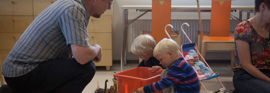 Kaksi pikkupoikkaa istuu lattialla leikkimässä, ja heidän vasemmalla puolella on mieshenkilö kyykyssä katsellen heitä ja oikealla puolella nainen istumassa lattialla.