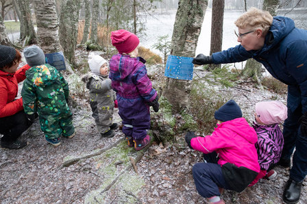 Ryhmä pieniä lapsia ja kaksi aikuista tutkii tehtävälappua marraskuisessa säässä metsässä järven rannalla.