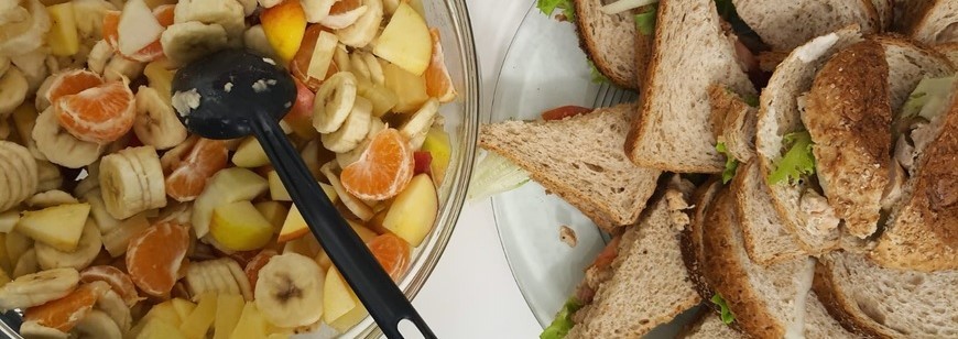 Kuvassa on hedelmäsalaattia kulhossa, jossa on kauha pystyssä sekä lautasella kolmioleipiä.