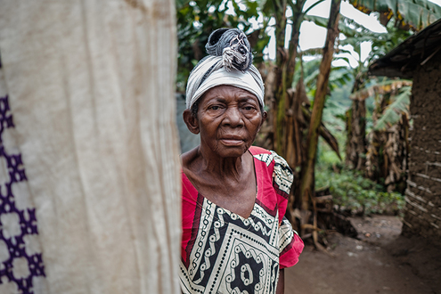 Kongolais-nainen seisoo pihalla kylässään. Hänellä on päässään huivi ja yllä kirjava mekko.