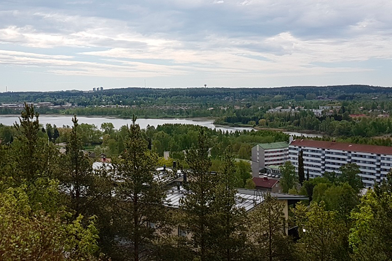 Kaukana alhaalla päin näkyy järvi jota ympäröi metsäinen maisema, vihreän metsän väleistä näkyy kerrostaloja.