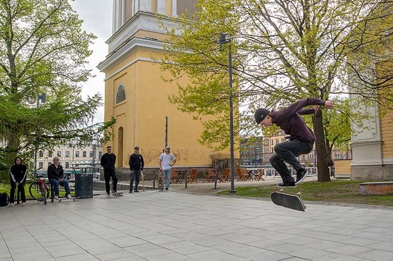 Poikaa skeittaa rullaulaudalla keltaisen puukirkon kirkkopuistossa, tekee hypyn ilmassa. Kaverit katsovat taustalla.