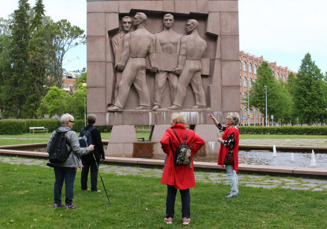 Kaksi punaliivistä naista kuvassa oikealla esittelee kahdelle kuulijalle suurta punakivistä patsasta joka on vesialtaan keskellä.