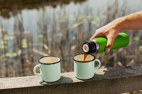 Aurinkoisella järven rannalla on parrun päällä kaksi emalimukia, joista toiseen joku kaataa kahvia termospullosta.