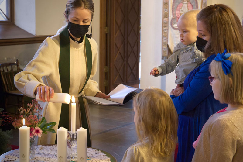 Pappi sytyttää pöydällä olevia kastekynttilöitä, ja kasteperhe seisoo pöydän oikella puolella katsomassa kynttilöiden sytyttämistä.