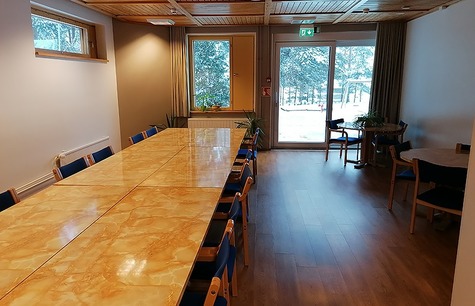 Tilakuva, jossa pitkä pöytä vasemmalla, jonka ääressä sinisiä tuoleja. Huoneen takana lasinen liukuovi joka johtaa ulos.