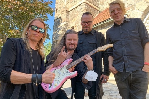 Neljä miestä Tuomiokirkon kesäisellä pihamaalla vaaleanpunaisen kitaran kanssa.