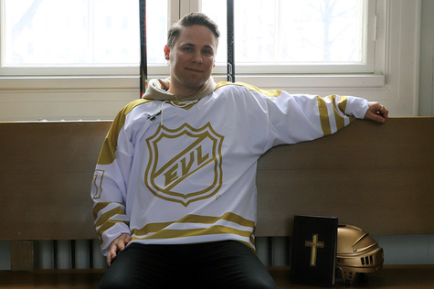 Valkoiseen jääkiekkopaitaan, jossa lukee EVL, pukeutunut mies istuu penkillä vieressään Raamattu ja kultainen kypärä.