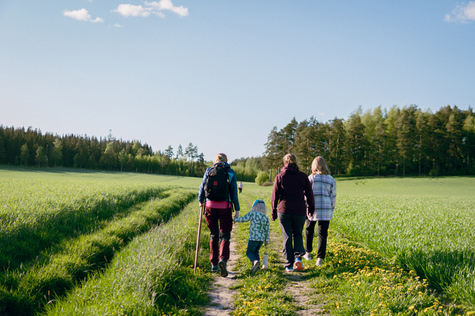 Aikuisia ja lapsi vaeltamassa suomalaisessa maalaismaisemassa.