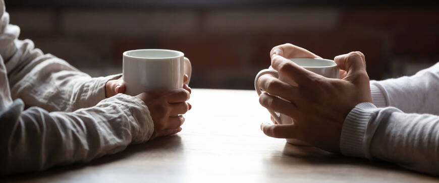 Pöydän ääressä vastakkain istuvista henkilöistä näkyvät kädet joissa on valkoiset kahvikupit.