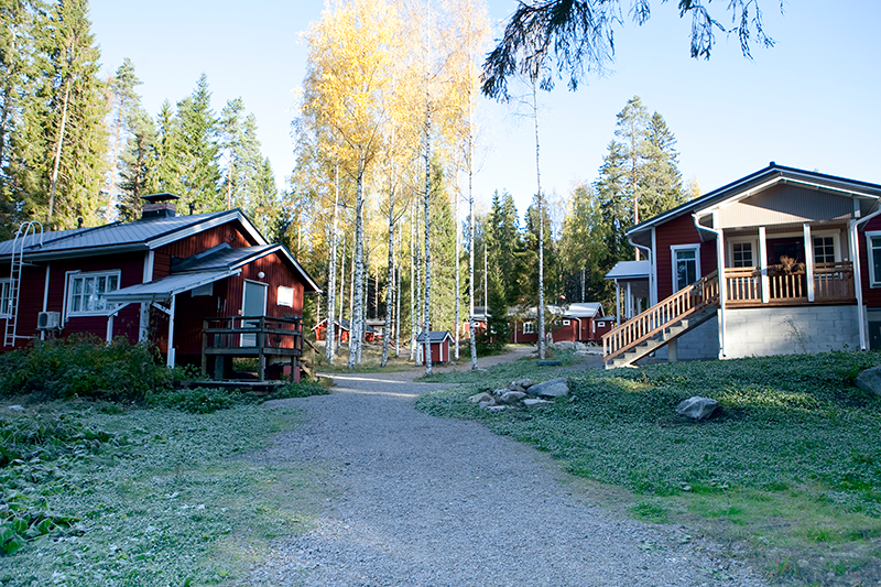 Kaksi punaista puutaloa hiekkapihan molemmin puolin metsän keskellä.