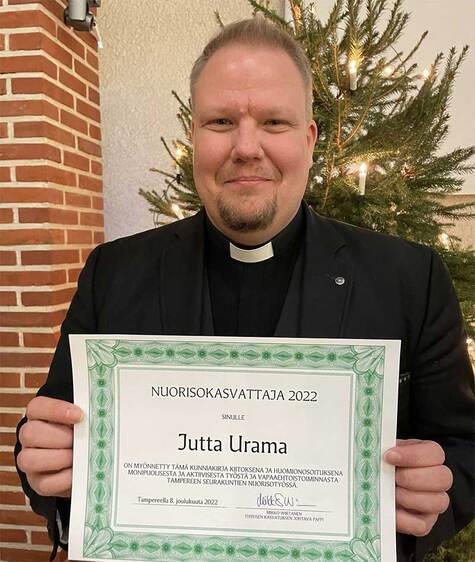 Papin puvussa oleva mieshenkilö pitää käsissä kuniakirjaa , jossa lukee Nuorisokasvattaja 2022 Jutta Urama.