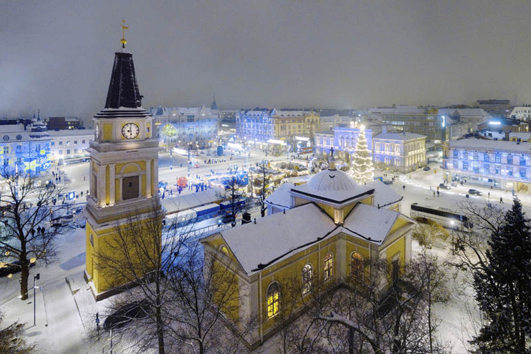 Vanha kirkko talvella yövalaistuksessa, taustalla joulutoria ja kaupungin joulukuusi.