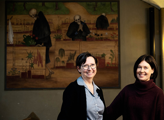 Kaksi naista nauraa taustallaan taulu, jossa luurangot kastelevat kukkia.