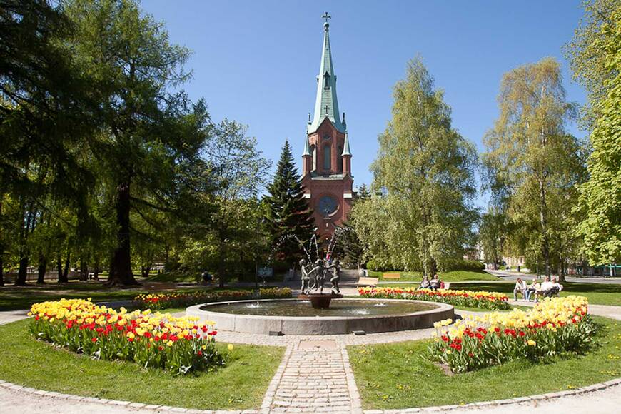 Pyynikin kirkkopuiston suihkulähde kevätpäivänä ja taustalla näkyy punatiilinen goottilainen kirkko.