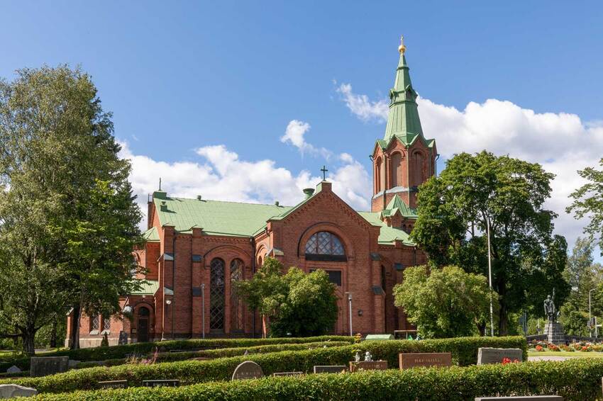 An overview of Messukylä Church, which is a front-facing red brick church in Neo-Gothic style, surrounded by green deciduous trees, with gravestones in the foreground.