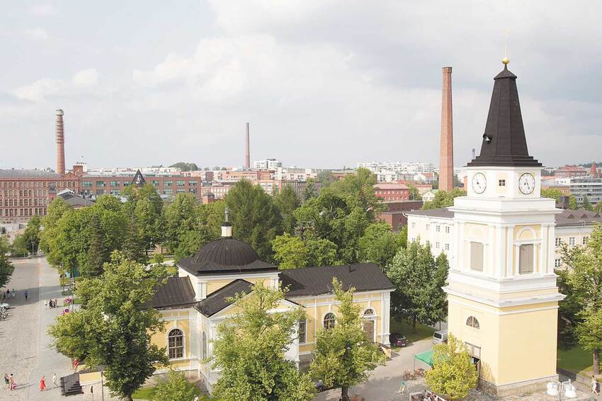 A summery aerial view of the light yellow wooden Old Church from the direction of the Central Square, with the bell tower to the right of the church, in the background green deciduous trees, and the red brick factory buildings of the Finlayson and Tampella areas with their chimneys.