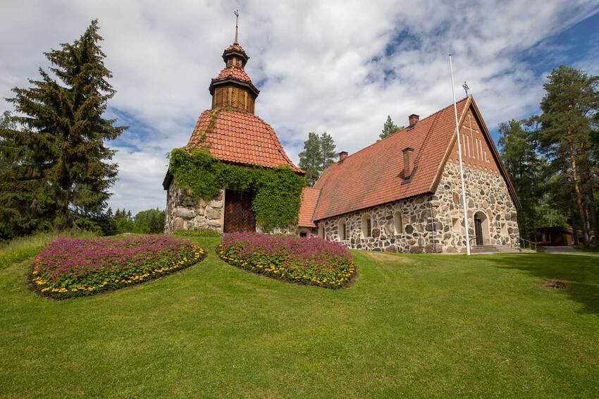 The old grey stone church, with a red tiled roof, sits atop a hill, and in front of the church, there is a green grassy area with flower beds.