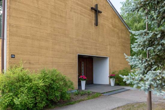 Vaalean ja matalan puisen kirkkorakennuksen tummien ovien yläpuolella on ruskeapuinen risti.