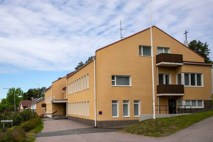 Keltainen suorakaiteen muotoinen matala rakennus on kuvattu rakennuksen päädystä, rakennuksen edessä on asfaltoitu piha ja muutamia vihreitä pensaita.