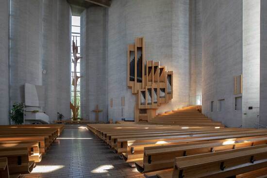 Harmaan betonisessa korkeassa kirkkotilassa  käytävän molemmilla puolilla on vaaleat puiset penkit ja käytävän päässä on korkea kapea ikkuna, jonka edessä on puinen korkea veistos.