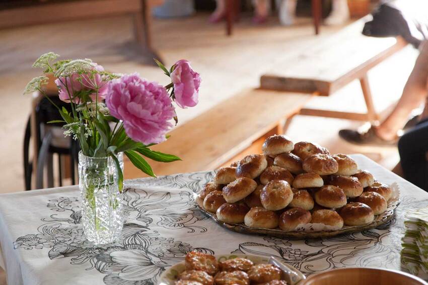 Vaalella pöytäliinalla peitetyllä pöydällä on kukkakimppu lasimaljakossa ja lautanen, jossa on keko pyöreitä pullia.