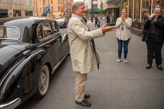 Hymyilevä mies vaaleassa popiilinitakissa seisoo musta vanhan auton vieressä, ja hänen ympärillään on ihmisiä, jotka taputtavat hänelle.
