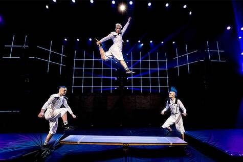 Kolme sirkustaitelijaa tekevät temppuja. Yksi taitelijoista hypännyt trampoliinilta korkealle ja kaksi taitelijaa odottavat vuoroaan patjoilla.