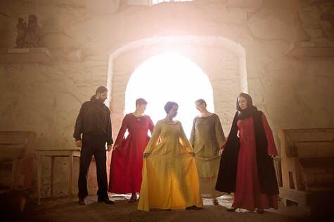 Yksi mies ja neljä kesiaikaisiin vaatteisiin puketunutta naista seisovat keskiaikaisessa kirkossa ja valo loistaa heidän takaansa olevasta oviaukosta.