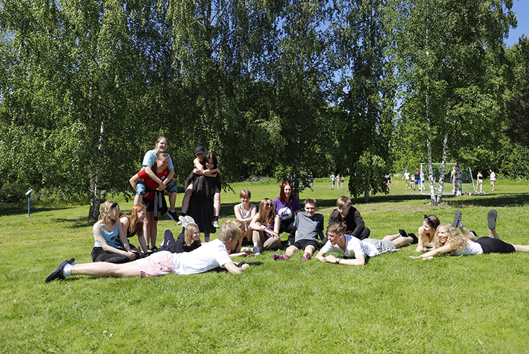 Nuoria makoilemassa nurmikolla kesällä. Osa nuorista pitää tosiaan reppuselässä.