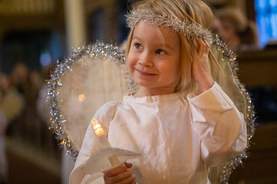 Onnellisen näköinen pieni lapsi on pukeutunut enkeliksi, ja hänellä on yllään valkoinen puku ja päässään sädekehä, hänellä kädessään kynttilää ja hänen taakseen on kiinnitetty hopeanhohtoiset siivet.