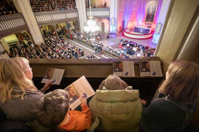 Ryhmä ihmisiä istuvat kirkon parvella, jossa on puisia koristeita, ja he katsovat alttarille ja pitävät ohjelmia käsisään.