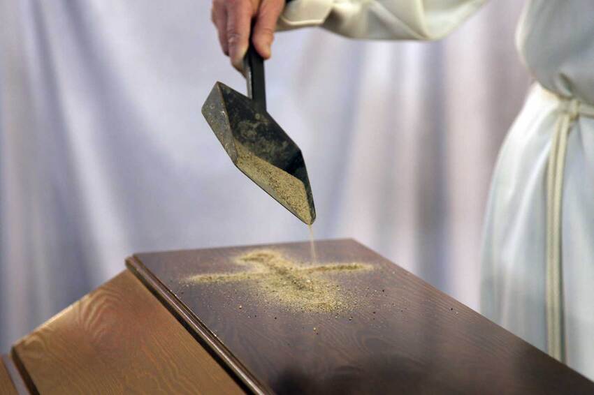 With a small shovel, sand is poured onto the top of the brown wooden coffin.