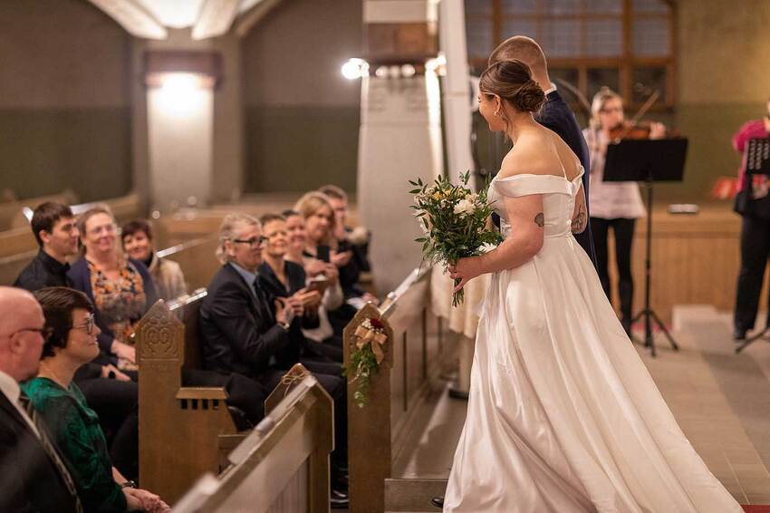 The bride, wearing a white dress and holding a bouquet of flowers, walks away from the altar, and wedding guests are seated in pews on both sides of the aisle.