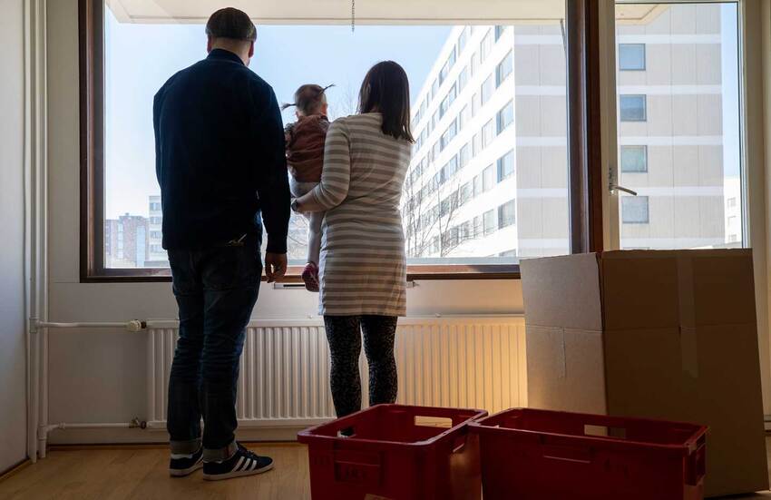 Tyhjässä huoneessa muuttolaatikoiden keskellä mies ja nainen, jolla on pieni lapsi sylissään katselevat isosta ikkunasta ulos, josta näkyy kerrostaloja