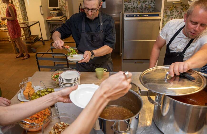 Pöydällä on kaksi isoa kattilaa, joista toisesesta otetaan keittoa lautaselle, pöydän toisella puolella seisoo kaksi kokkia, joista toinen laittaa salaatia lautaselle.