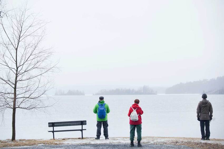 Kolme ulkoiluvaatteisiin pukeutunutta ihmistä seisovat jäätyneen järven rannalla ja katsovat lumiselle ulapalle.