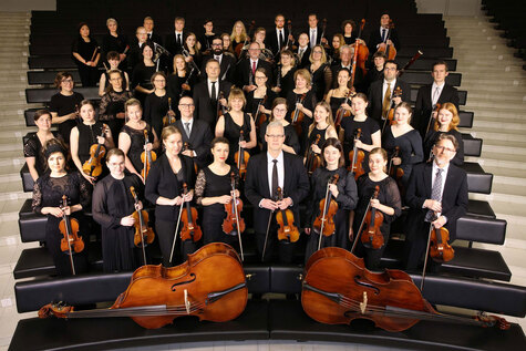 Mustiin pukeutuneut sinfoniaorkesteri seisoo portaikoissa  ja heilla on jousisoittimeia käsissään.